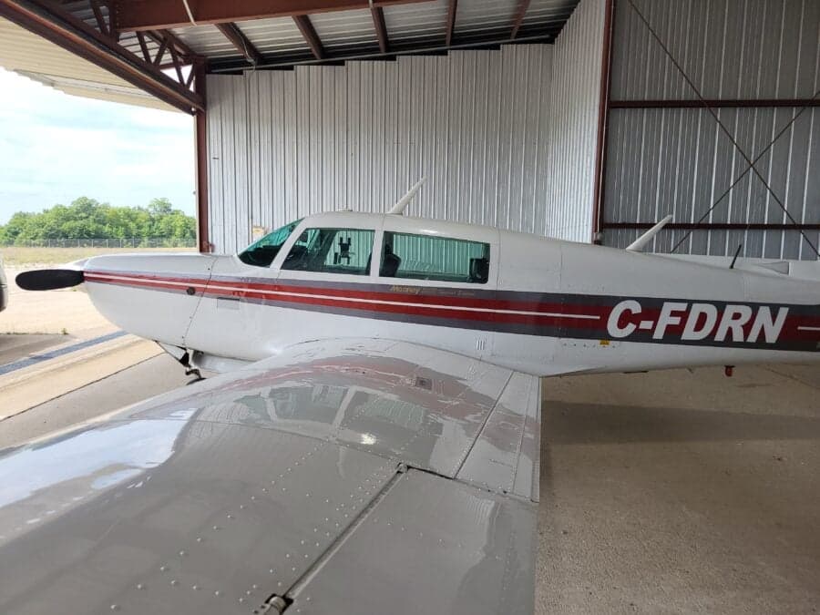 A side view of a white Mooney M20J aircraft with red and grey stripes, registration C-FDRN, parked inside a hangar.