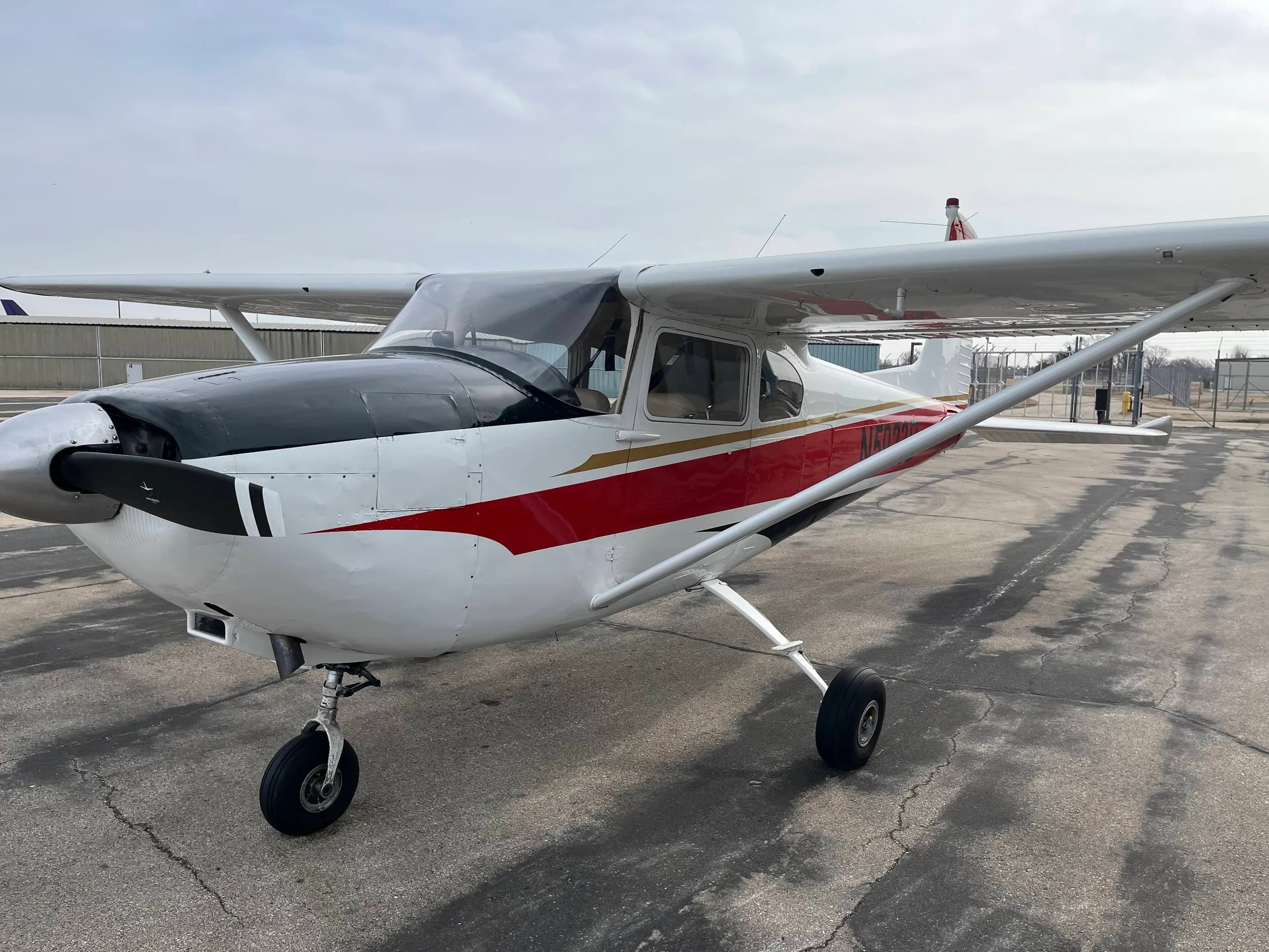 A three-quarter front view of a white Cessna 182 aircraft with red and black stripes, parked on an asphalt apron.