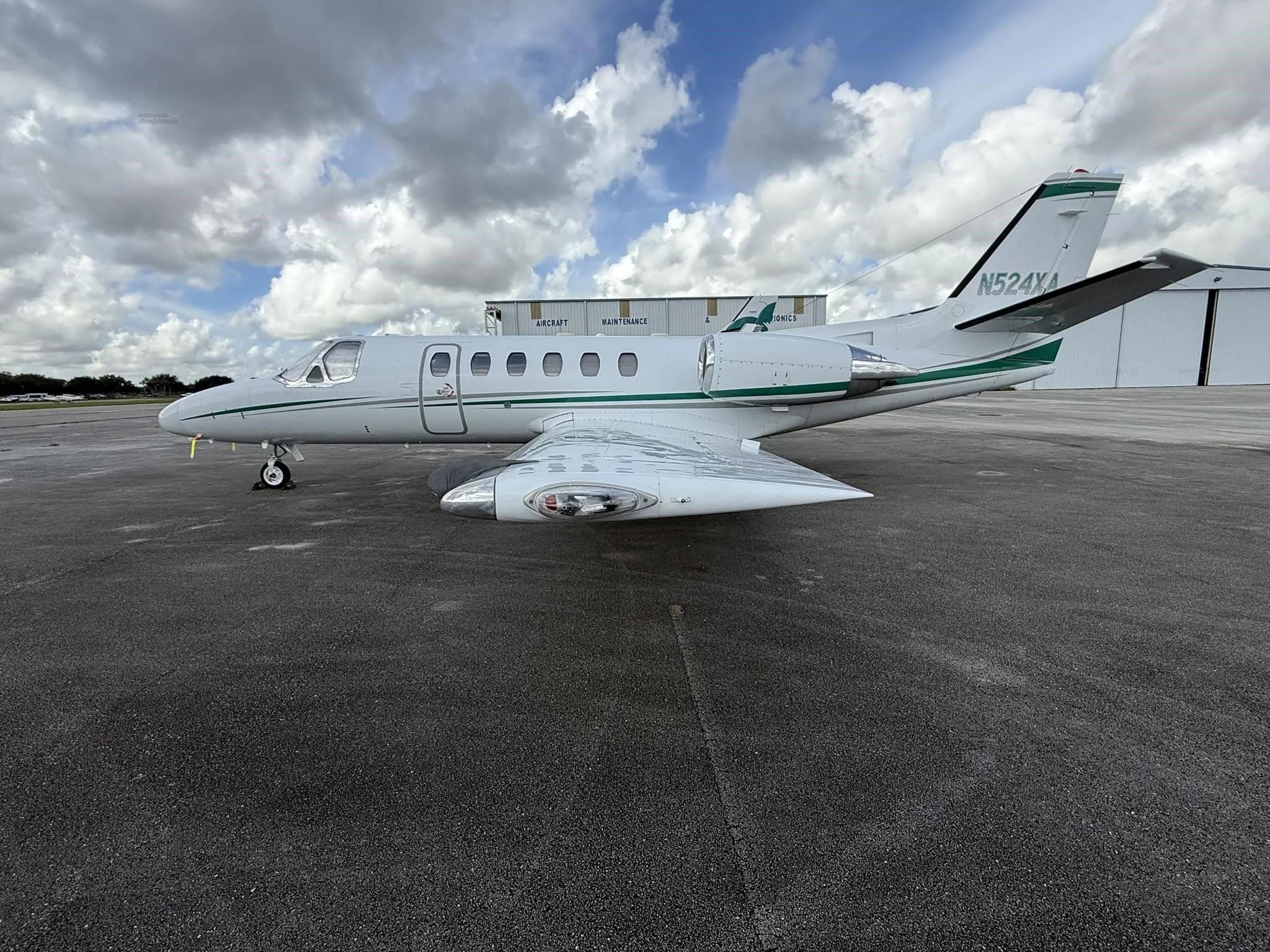 A white business jet N524XA with green stripes, parked on an airport tarmac with hangars in the background.