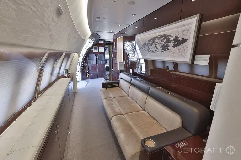 A long view of a luxurious aircraft cabin with a beige sofa, dark wood accents, and artwork on the wall.