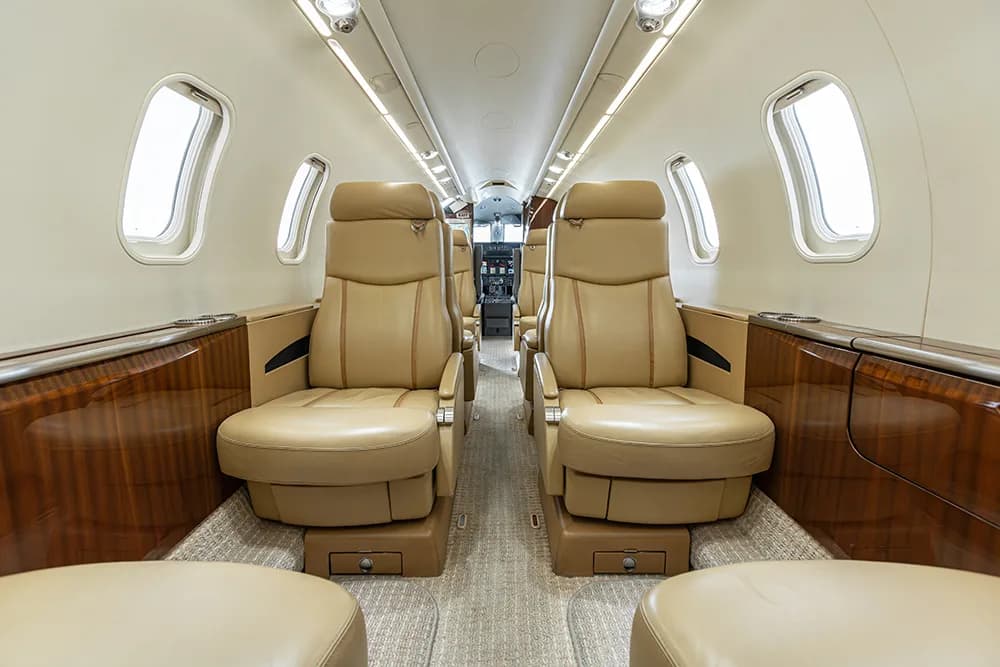A view down the private jet cabin aisle towards the cockpit, showing tan leather seats and wood paneling.
