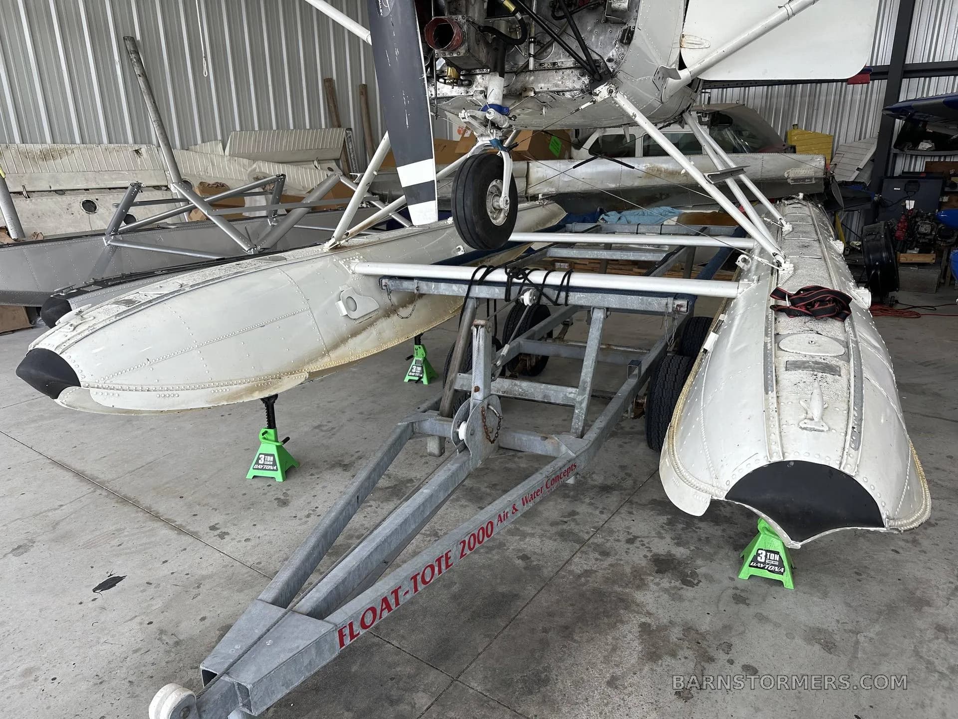 A white floatplane with its propeller visible is on jacks in a hangar, showing the floats and a "FLOAT-TOTE 2000" trailer.