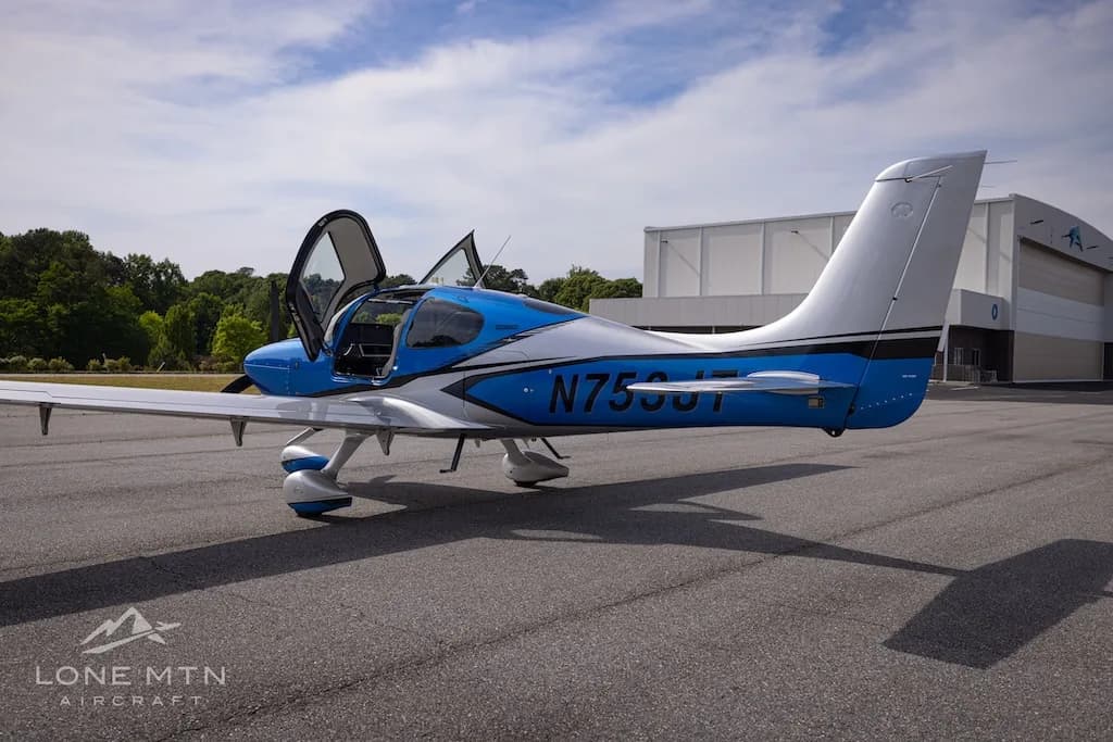 A blue and silver single-engine aircraft, N753JT, with open gull-wing doors is parked on an asphalt tarmac next to a hangar.