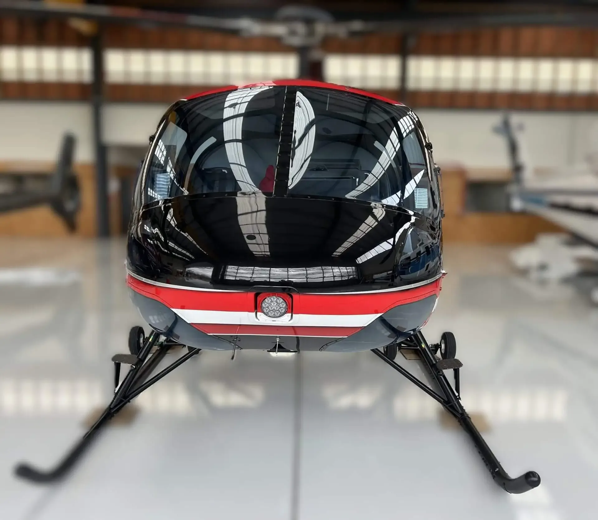 A rear view of the red and black helicopter inside a hangar, showing its tail and landing skids.