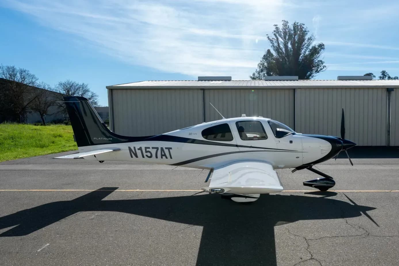 A side view of a white and dark blue Cirrus SR22 aircraft, registration N157AT, parked on an asphalt apron with a hangar in the background.