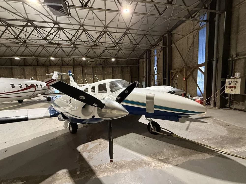 Front-right view of a white and blue Piper Seneca twin-engine propeller aircraft parked in a hangar.