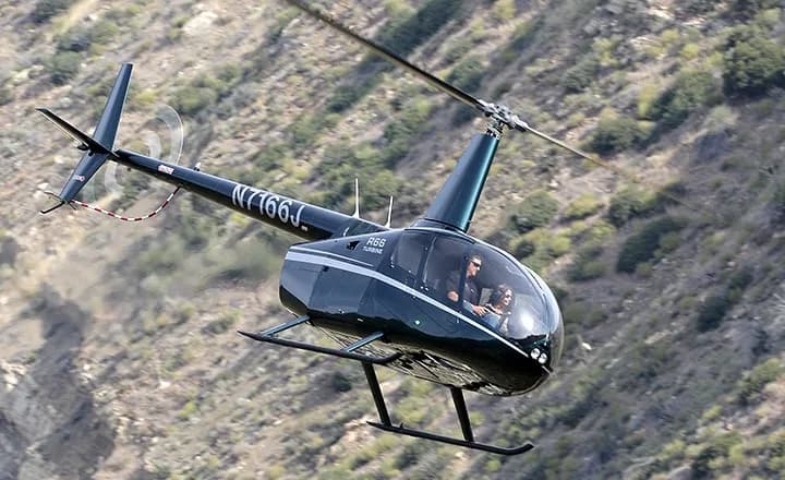 A dark green Robinson R66 Turbine helicopter in flight with two people inside.