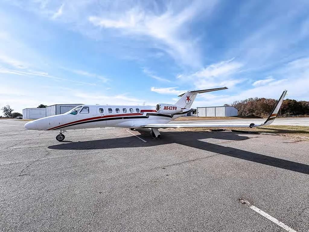 A white Cessna Citation private jet with red and black stripes and tail logo, parked on an asphalt tarmac with hangars in the distance.