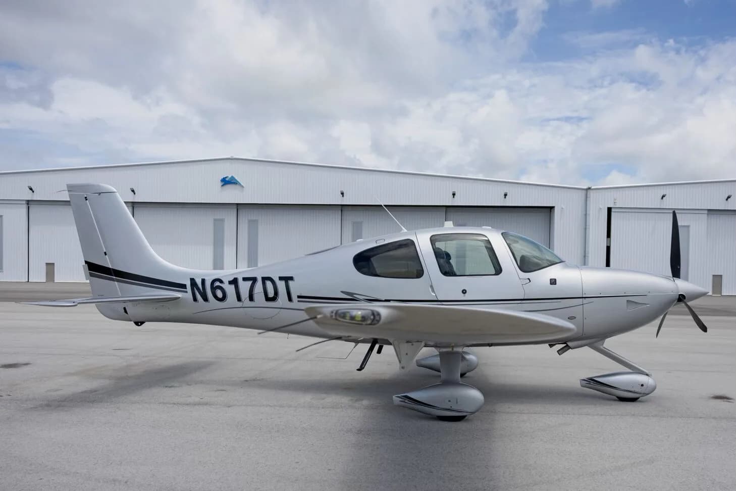 A silver Cirrus SR22 aircraft with tail number N617DT parked on an tarmac in front of hangars.