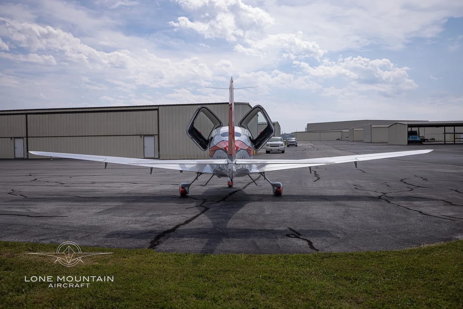 A rear view of a silver and red Cirrus SR22T aircraft with registration N211CK parked on an asphalt ramp with its gull-wing doors open.