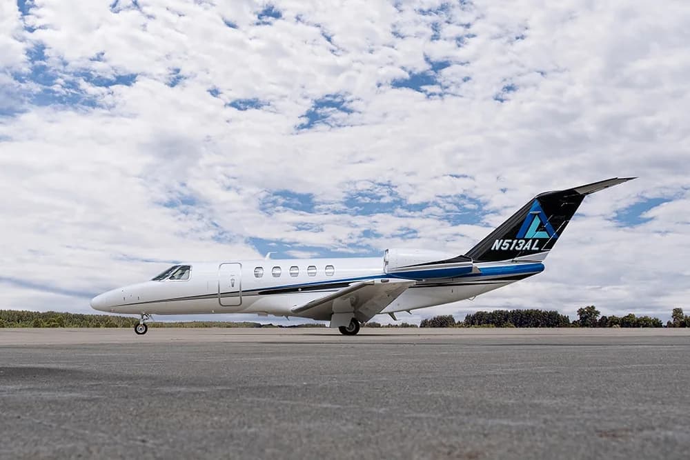 A white private jet with blue and black tail accents (N513AL) is parked on an airfield under a partly cloudy sky.