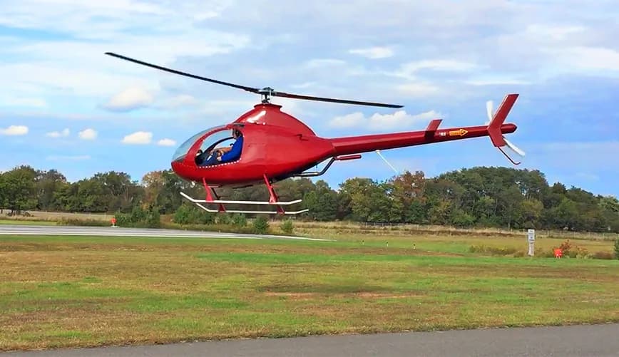 A red RotorWay Exec helicopter hovers over a grassy field near a runway.