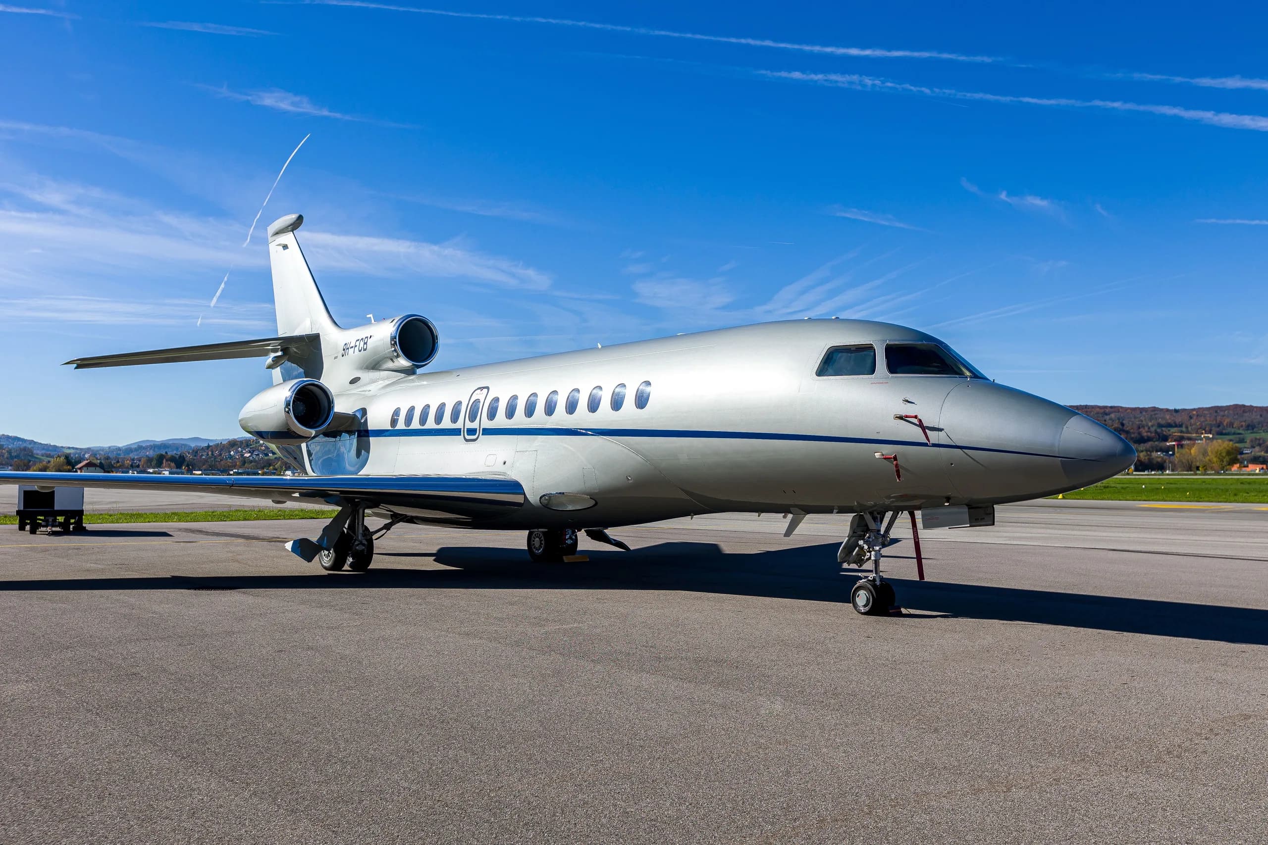 A silver Dassault Falcon 900EX business jet with blue stripes parked on an airfield, viewed from the front-right.