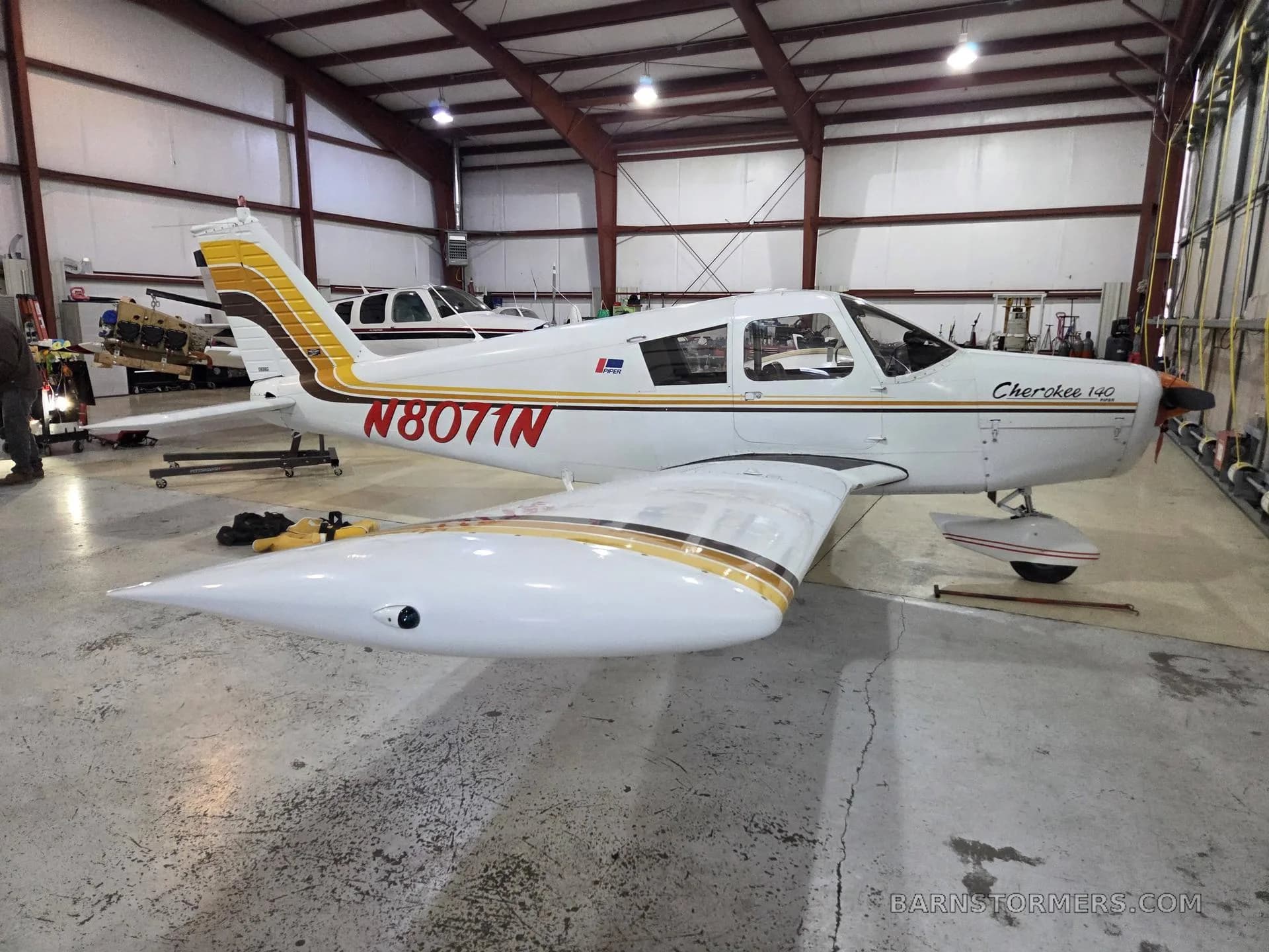 A white Piper Cherokee 140 with brown and gold stripes, registration N8071N, parked in a hangar.