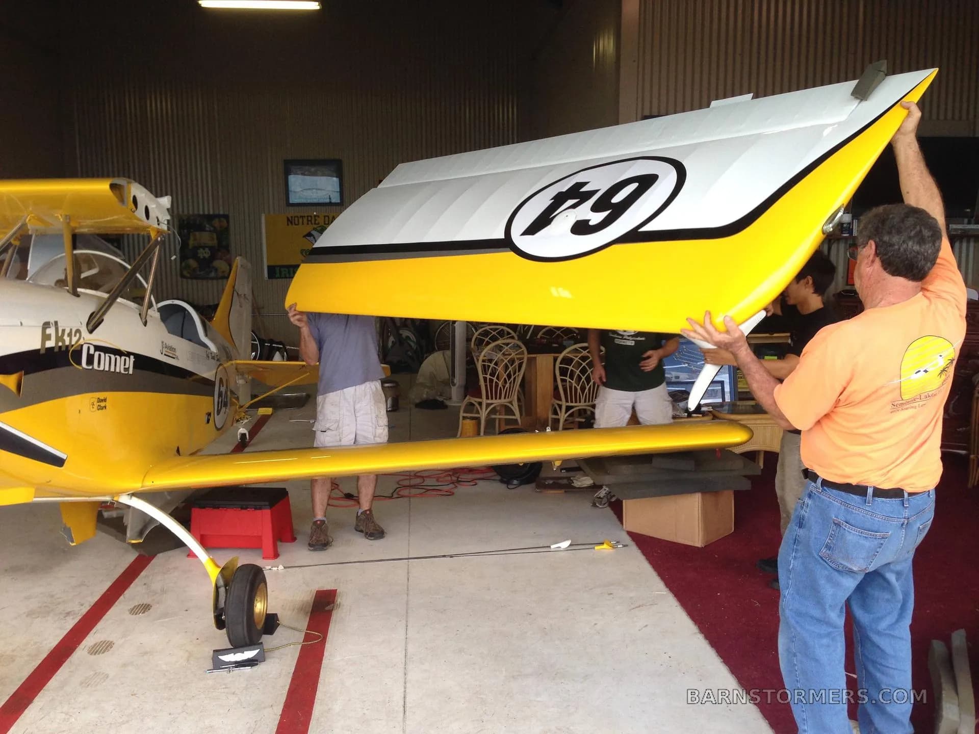 Two men working on the tail section of a yellow and white FK12 Comet biplane in a hangar.