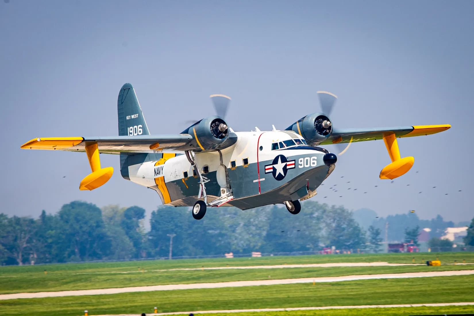 A Grumman HU-16 Albatross seaplane in US Navy livery with '906' on the nose, flying low over a green airfield with landing gear extended.