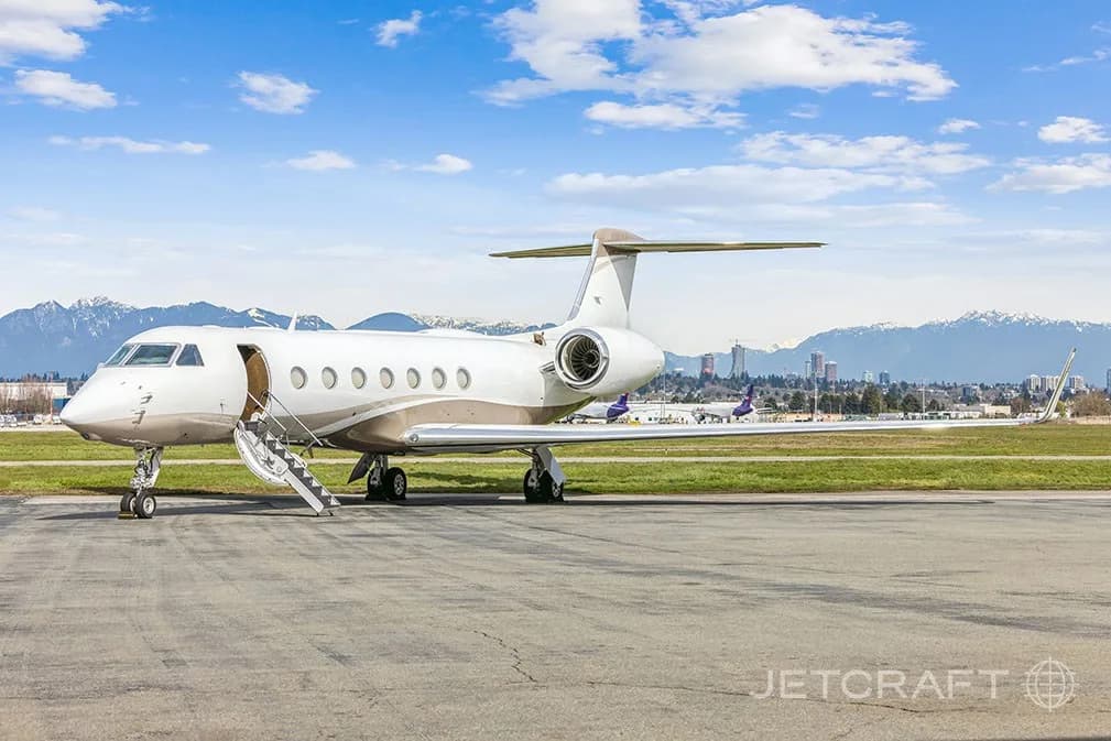 A white and tan Gulfstream G550 business jet parked on an airfield with mountains in the background.