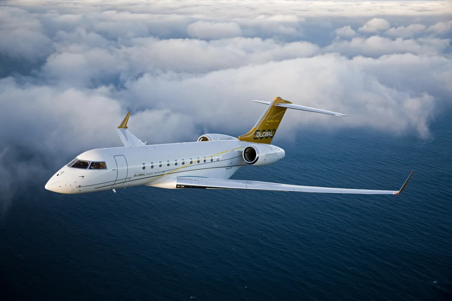 A white Bombardier Global 5000 business jet with gold tail accents flies above the ocean with clouds in the background.