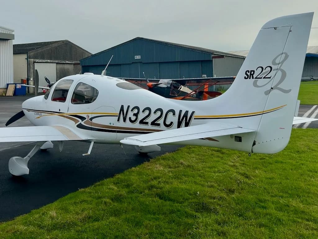 A rear-side view of a white Cirrus SR22 aircraft with N322CW registration, parked on a tarmac next to green grass.