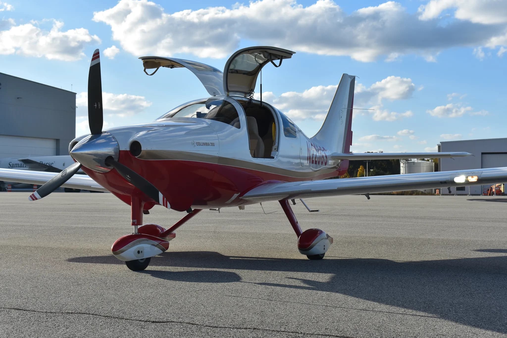 Front-quarter view of a white and red Columbia 350 aircraft with both gull-wing doors open, parked on an airfield.