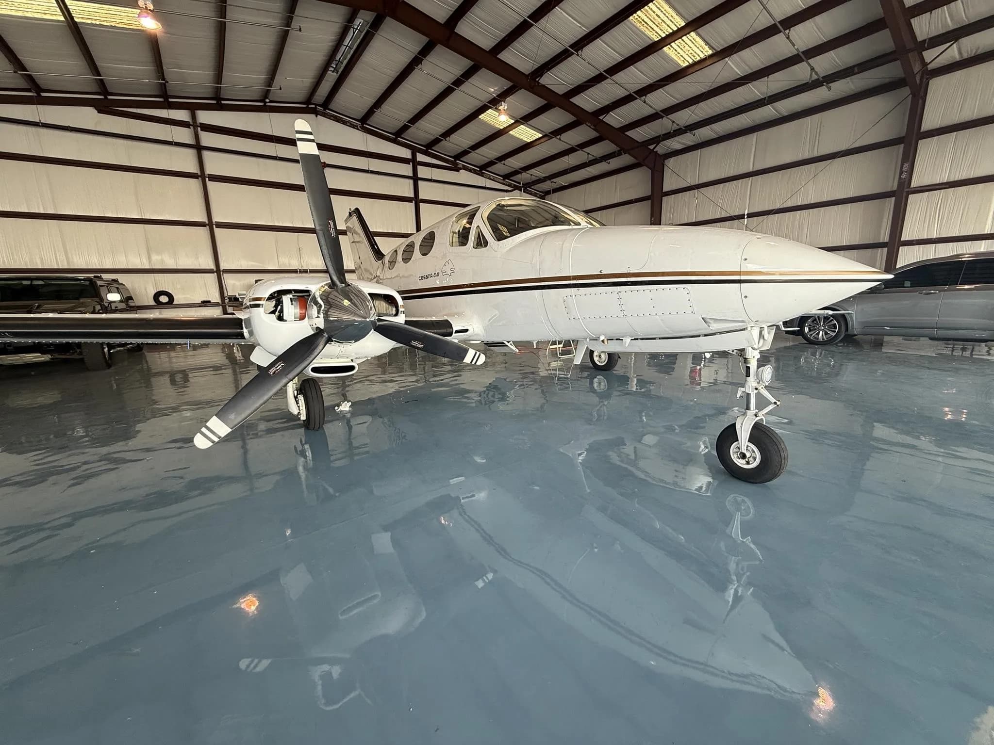 A white twin-engine turboprop aircraft with gold stripes is parked inside a hangar with a shiny gray floor.