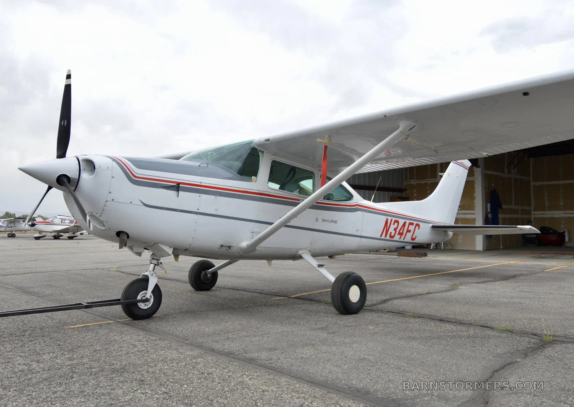 A front-left view of a white Cessna 182 Skylane with red and gray stripes, registration N34FC, parked on an asphalt ramp.