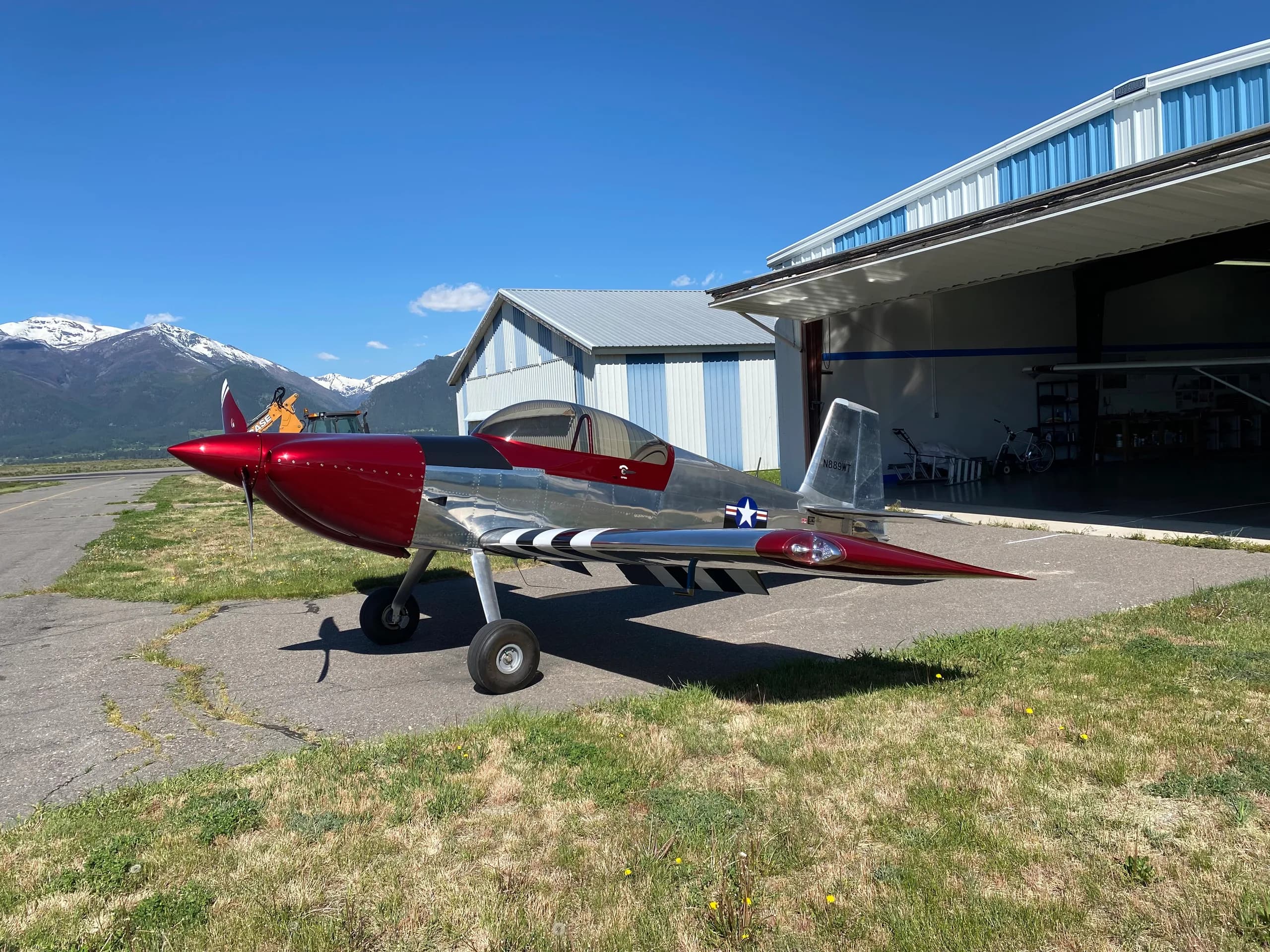 A shiny silver and red Vans RV-8 aircraft with black and white invasion stripes on the wings, parked outside a hangar with mountains in the background.