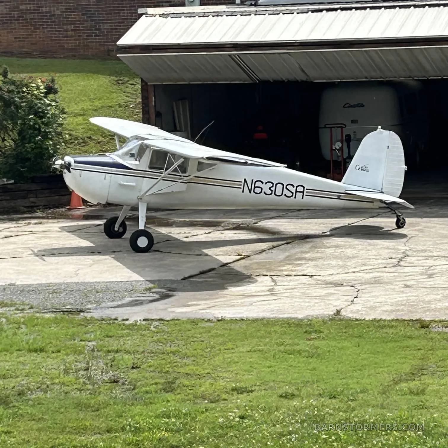A white Cessna 170B with dark blue and gold stripes is parked on a concrete pad outside a garage.