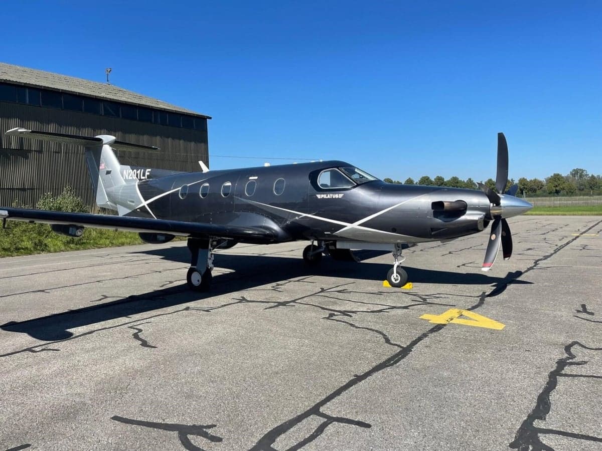A dark grey and silver Pilatus PC-12 turboprop aircraft with white accents is parked on an asphalt tarmac under a clear blue sky.
