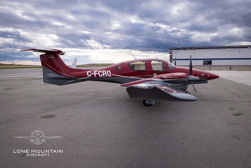 A red and grey Diamond DA62 aircraft with registration C-FCRQ parked on an airfield next to a hangar.