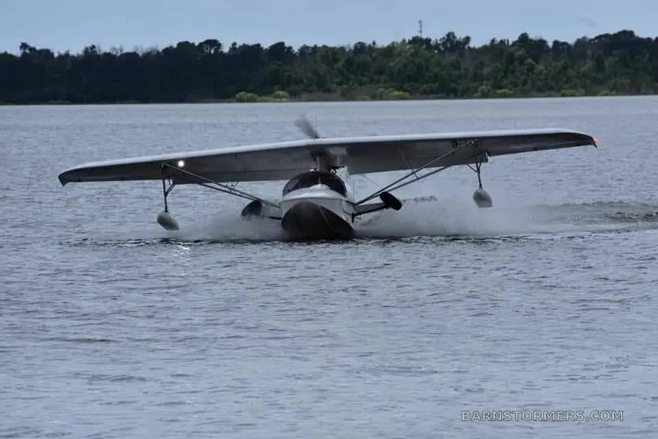 A white experimental seaplane is taking off or landing on water, creating a large splash.