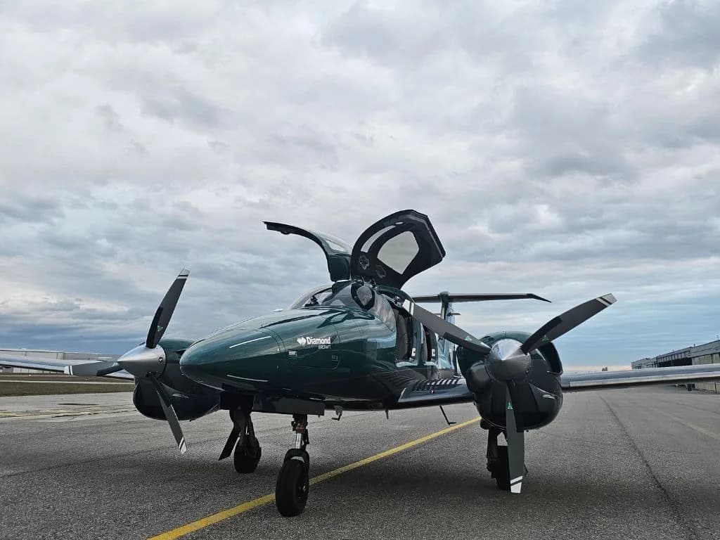 A dark green Diamond DA62 aircraft with its doors open, parked on a tarmac under a cloudy sky.