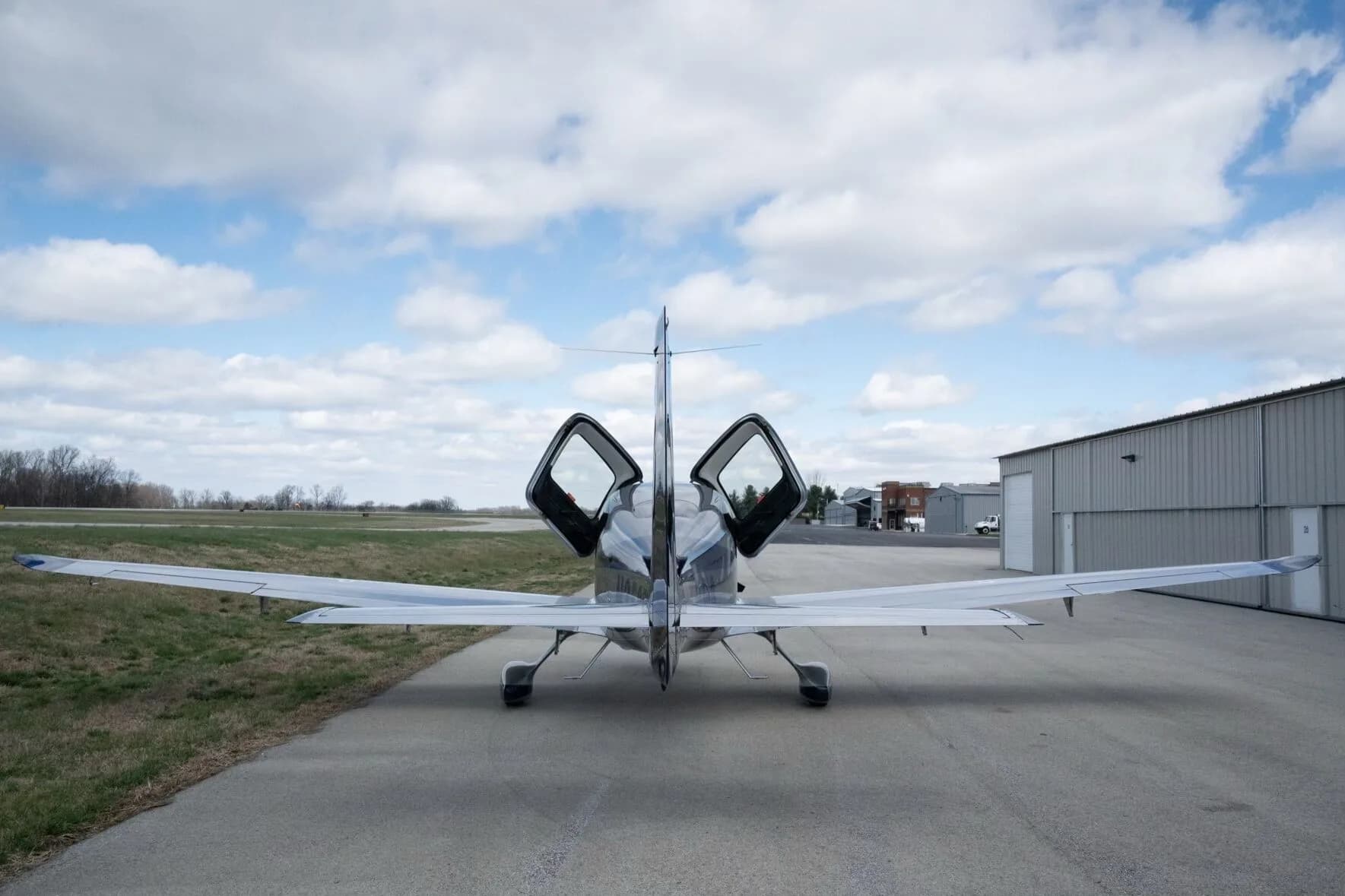 Rear view of a silver and blue Cirrus SR22T aircraft with its gull-wing doors open, parked on an airfield.