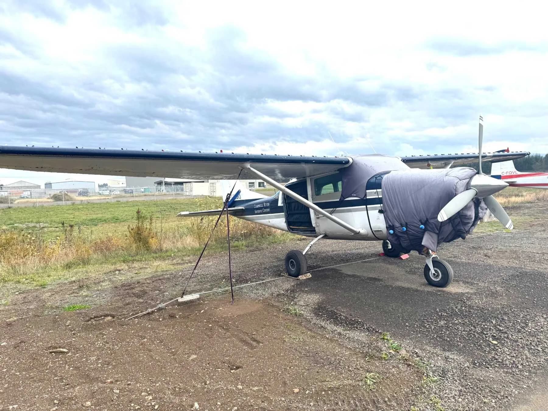 A Cessna 206 Stationair parked on a gravel area with the door open and a cover on the engine.