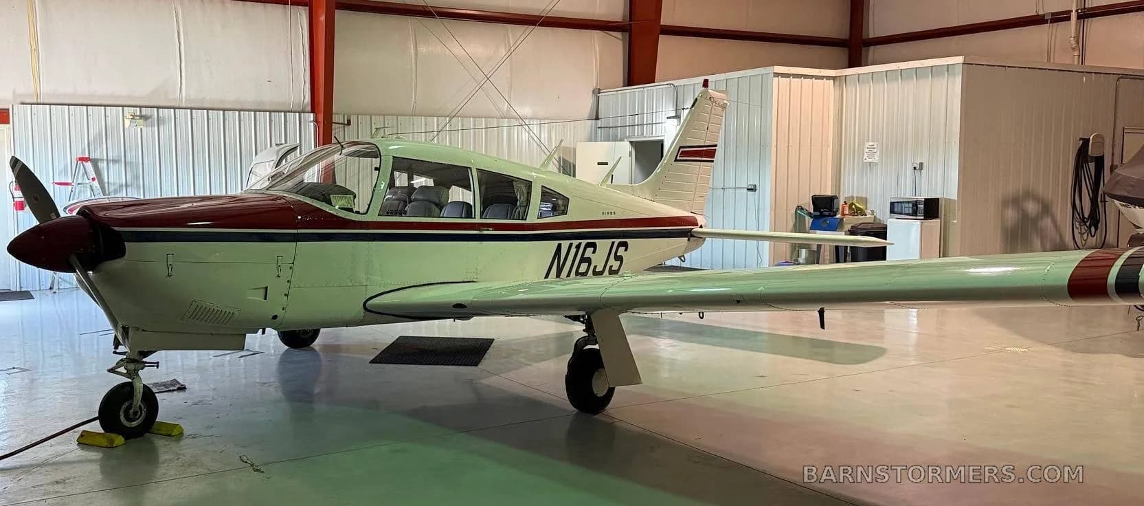 A white Piper aircraft with red and blue stripes, registration N16JS, parked inside a hangar.