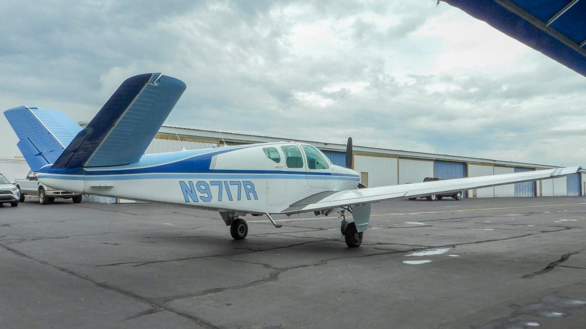A white and blue Beechcraft Bonanza parked on the tarmac with hangar in the background.