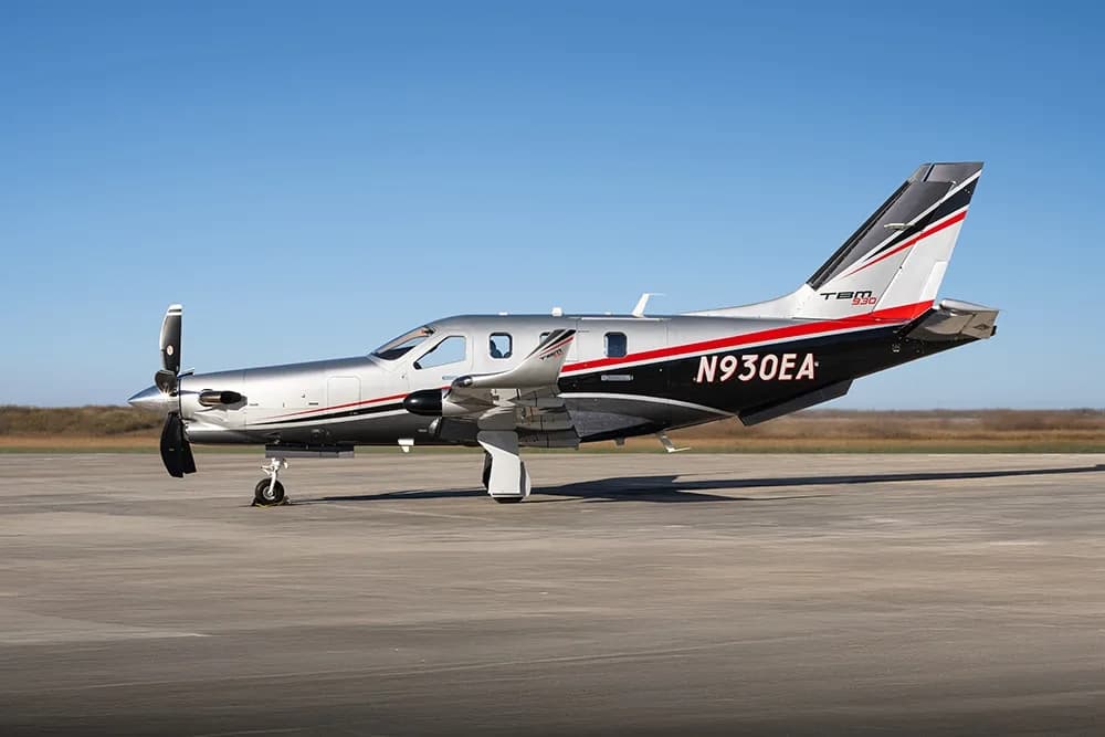 A silver and black Daher TBM 930 turboprop aircraft with red and white accents is parked on a tarmac under a clear blue sky.