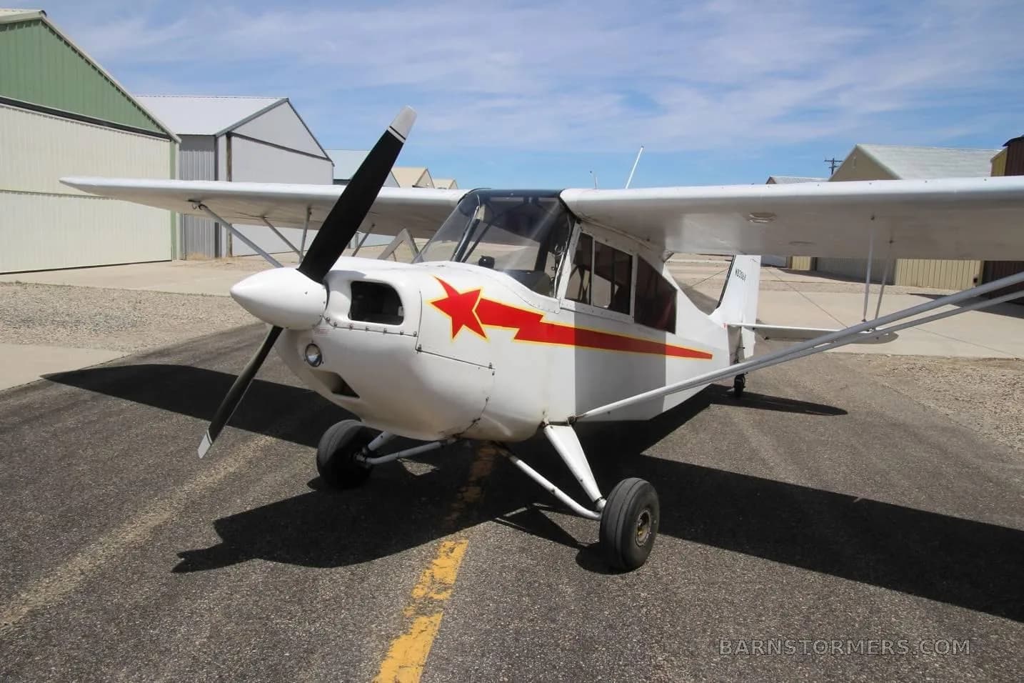 A front view of a white single-engine high-wing aircraft with a red star on the cowl and red stripes, parked on an asphalt taxiway.