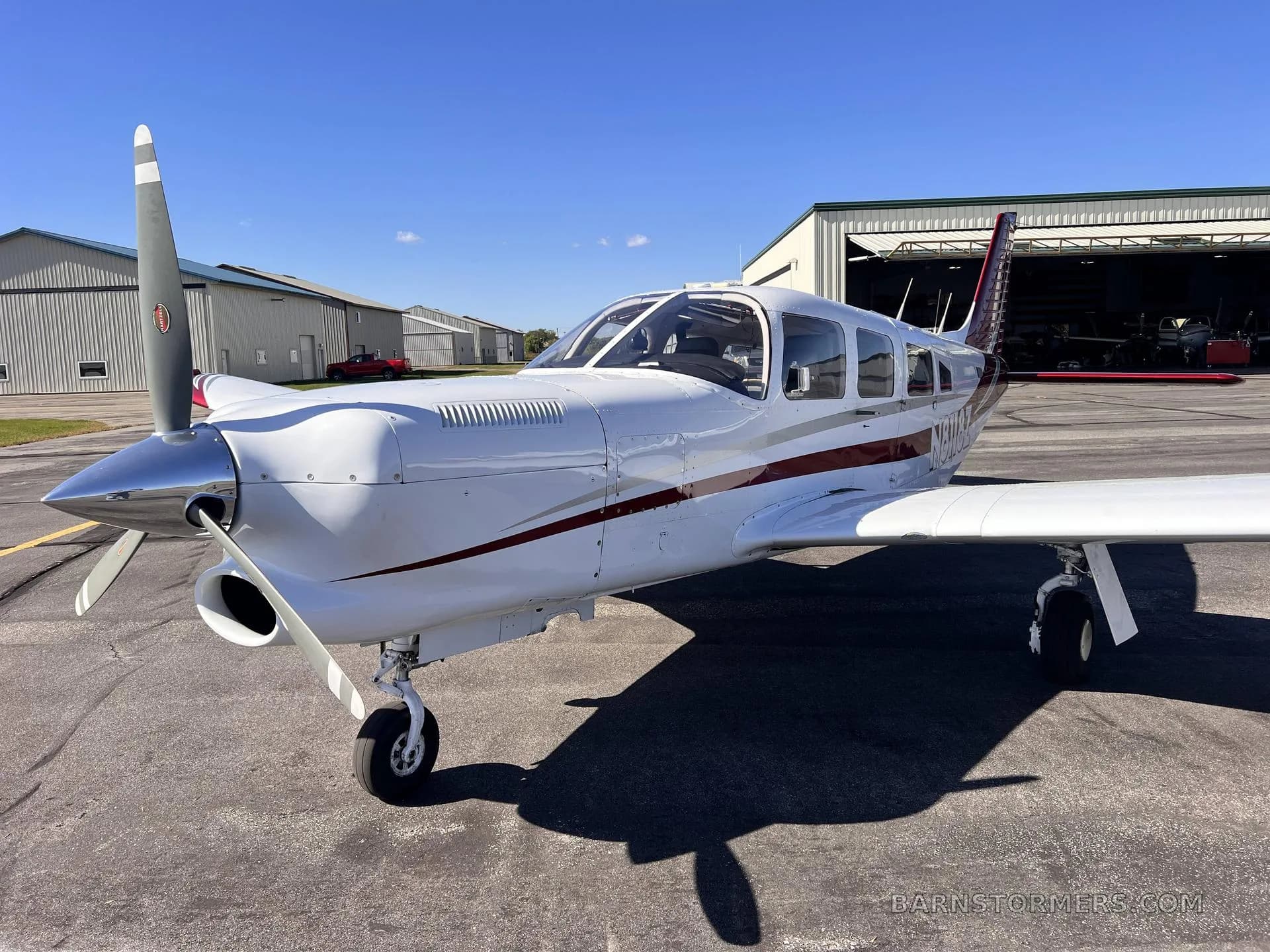 The front-right view of a white single-engine propeller aircraft with red stripes, parked on an asphalt ramp in front of hangars.