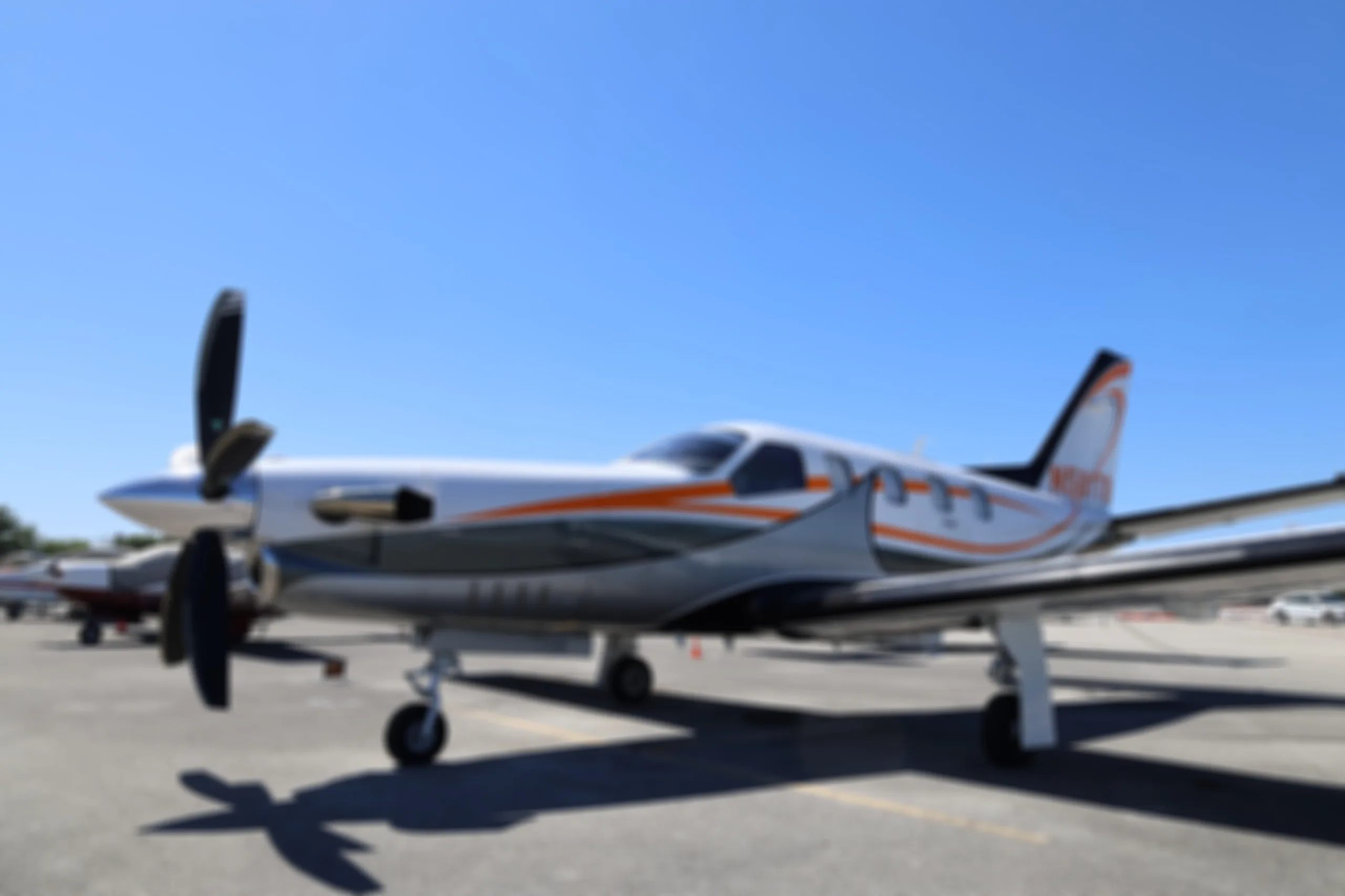A blurry photo of a silver aircraft with orange and black stripes parked on an airport tarmac under a blue sky.
