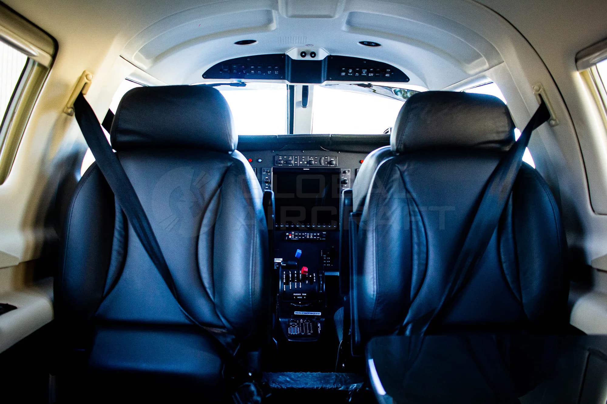 The interior cabin of an aircraft, showing two black leather seats with seatbelts, looking forward towards the cockpit.