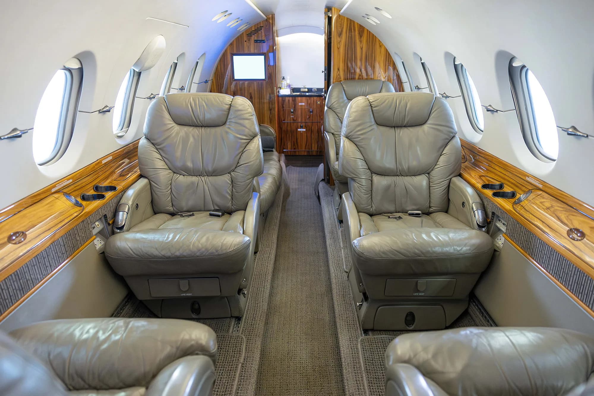The interior cabin of a business jet with grey leather seats and wood paneling, looking towards the rear galley area.