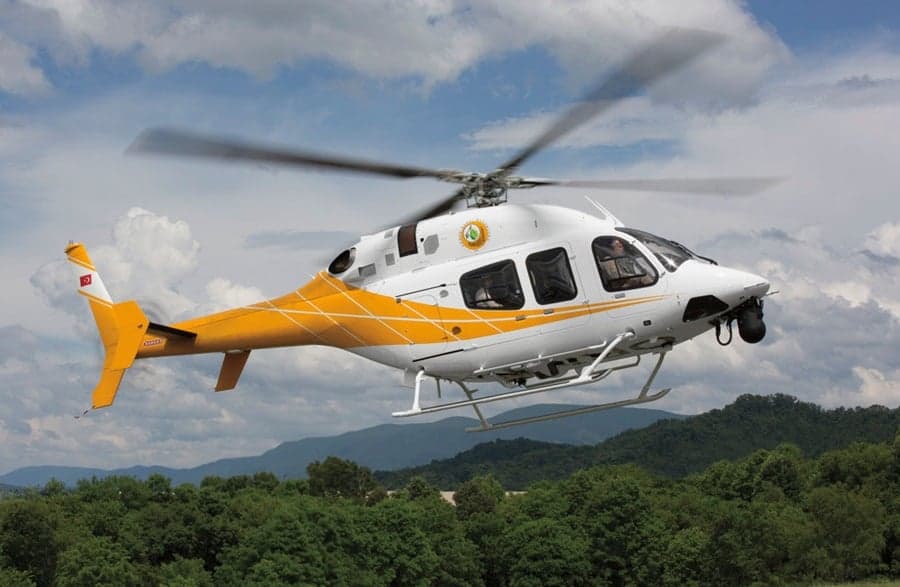 A white and yellow helicopter flies over a forest.
