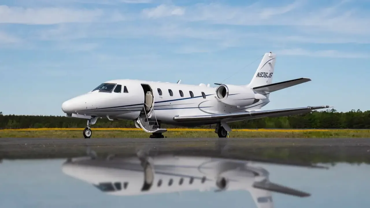 A white Cessna Citation XLS+ business jet, tail number N836JS, with blue stripes, parked on a tarmac with its main door open, reflected in a puddle.