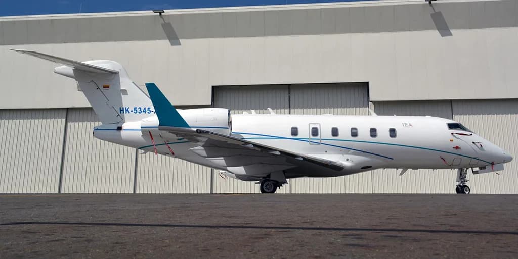 A white business jet with blue and teal stripes, registration "HK-5345-X" and "IEA", is parked on a tarmac in front of a hangar.