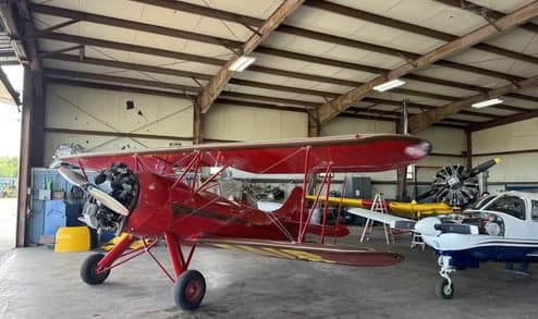 A red biplane with an exposed engine is parked in a hangar, with other aircraft and parts in the background.