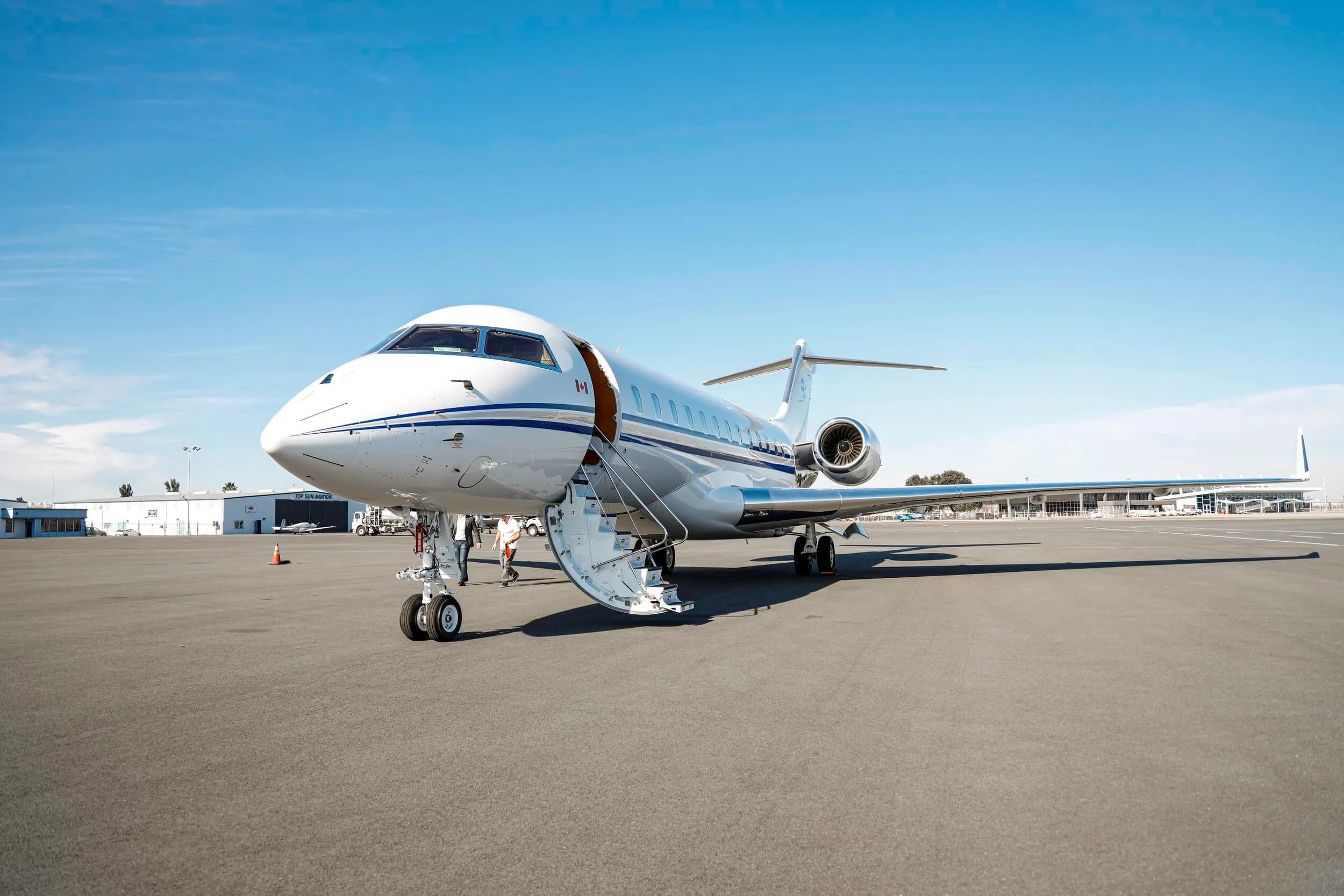 A white private jet with blue stripes is parked on an airport tarmac with its main door open and stairs extended.