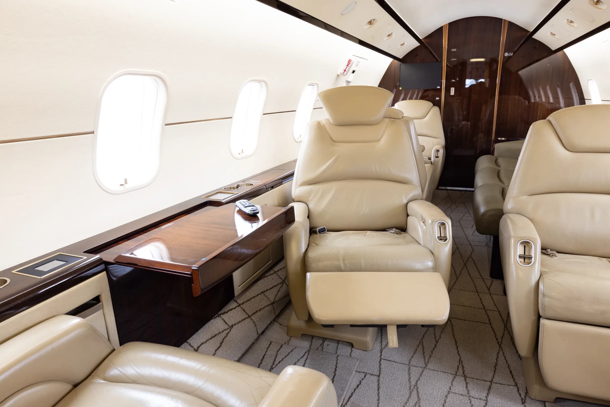 A close-up view of a light beige leather recliner seat and a polished wooden side table in a business jet cabin.
