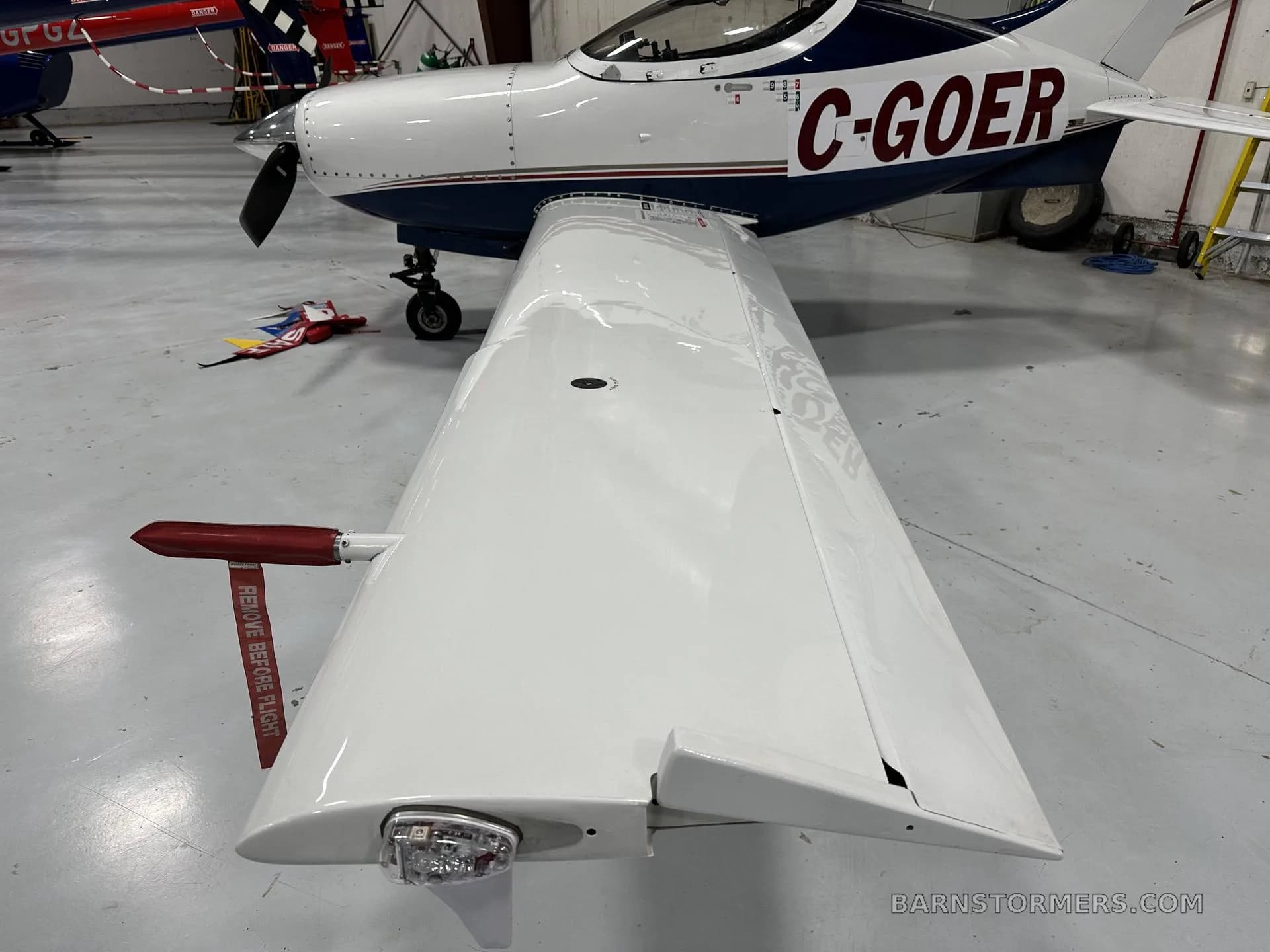 A close-up view of the right wing of a white and blue aircraft, showing the wingtip light and a "REMOVE BEFORE FLIGHT" tag, inside a hangar.