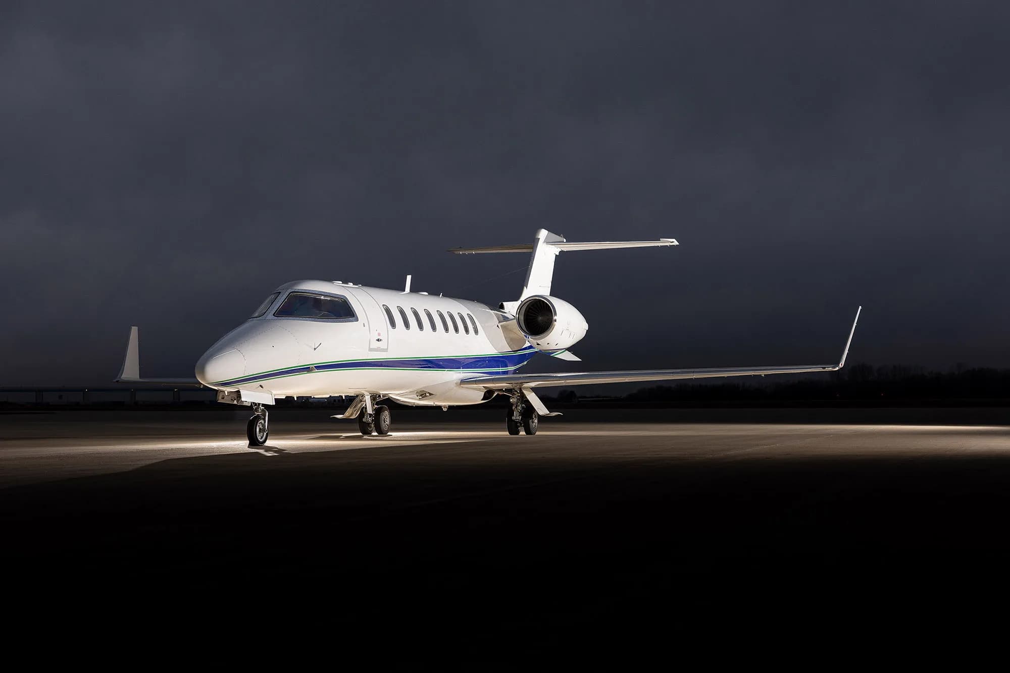 A white Learjet 75 with blue and green stripes parked on an tarmac under dramatic cloudy skies, viewed from the front.